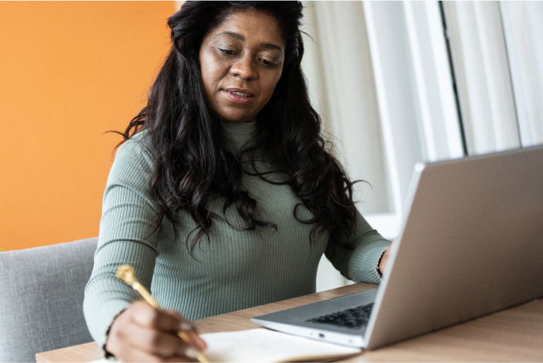 Black woman works on computer and writes in a notebook.