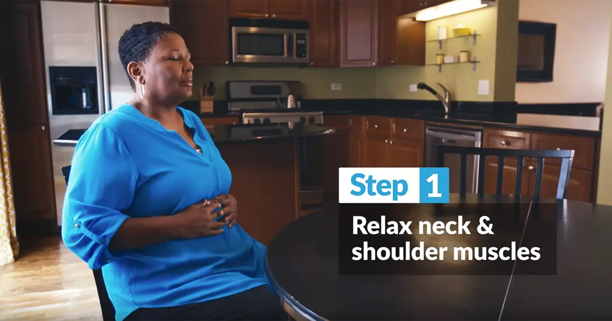 Woman sits in kitchen with hands on belly taking a deep breath.
