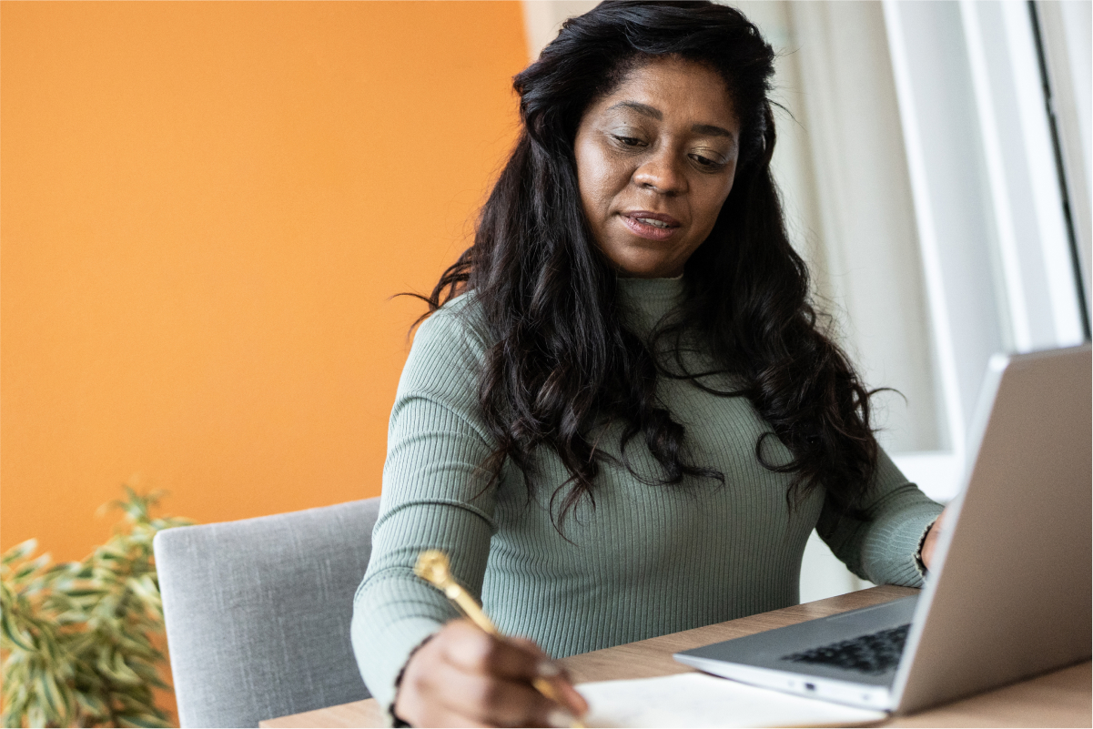 Black woman works on computer and writes in a notebook.
