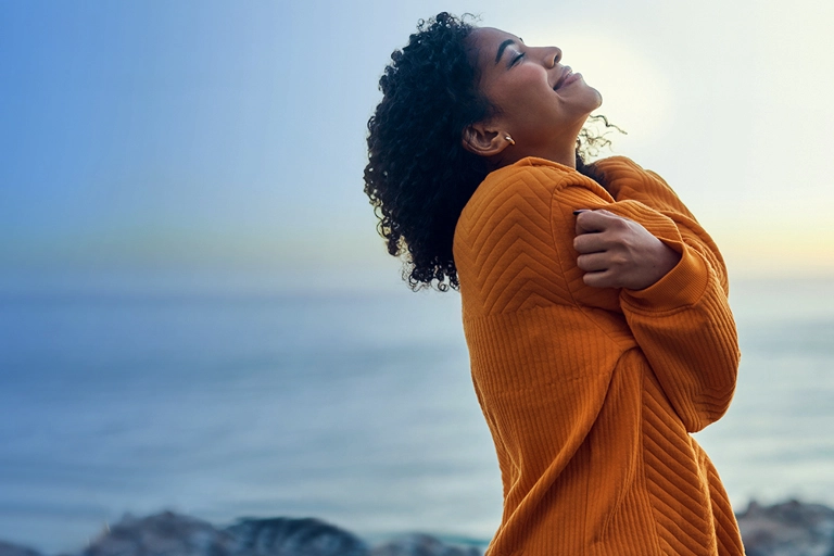 Woman in orange sweater hugs her arms and looks to the sky against coastal sunrise